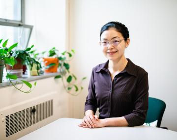 坐在一张桌上的亚裔女性用她的手横渡了。她戴着眼镜和紫色纽扣衬衫，并在辫子中捆绑着她的头发。她坐在一个白墙办公室。